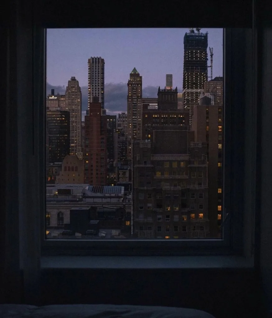 a city skyline seen through a bedroom window at blue hour, lit windows against a fading sky